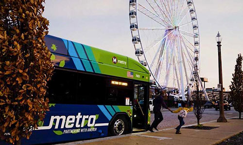 Electric bus sitting in front of the St. Louis Wheel with a father and daughter getting off of the bus, heading to the Wheel